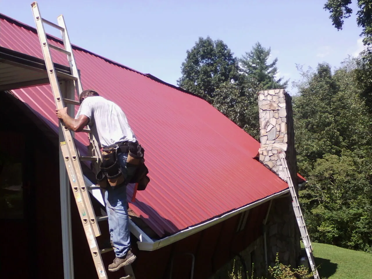 Roofer on a ladder beside a red metal roof and stone chimney — Soffit & Fascia Repair by Roofing Brothers in Elko